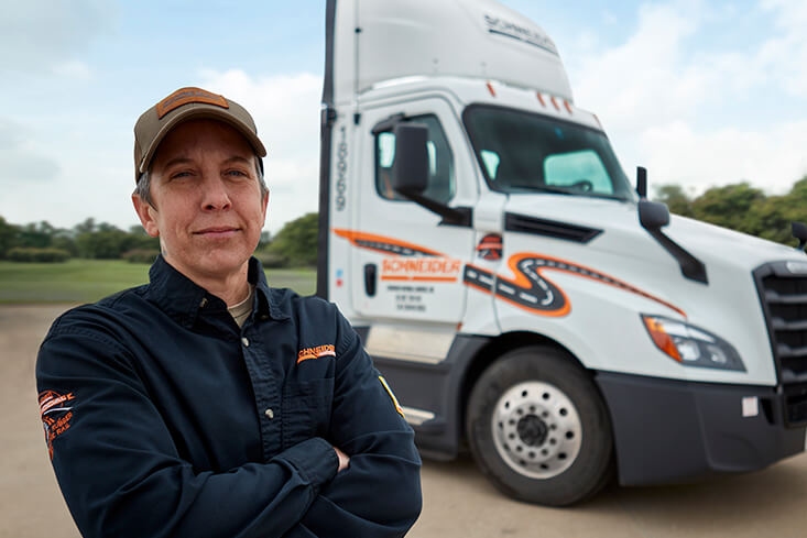 A local Schneider driver stands with their arms crossed in front of a white day cab tractor