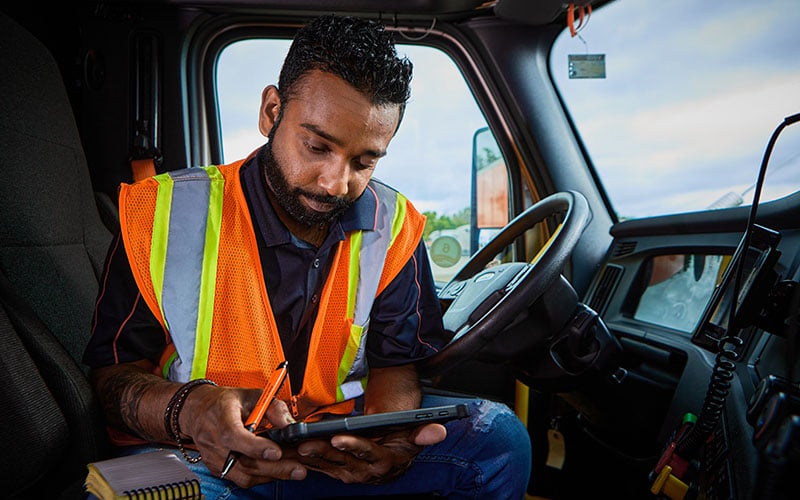 A Schneider driver in a hi-vis vest, sitting in the driver's seat of a parked semi-truck, working on his tablet.