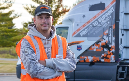 Ashton Frank standing in front of a military-themed semi-truck while wearing a Schneider hat and orange safety vest.