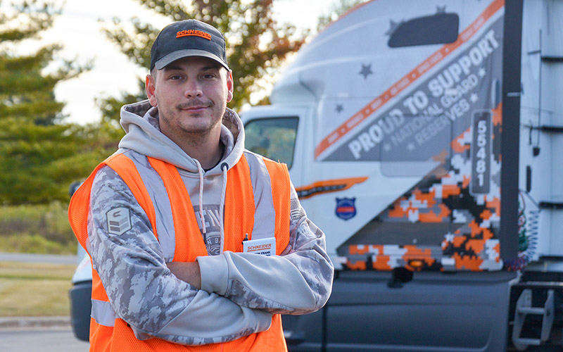 Ashton Frank standing in front of a military-themed semi-truck while wearing a Schneider hat and orange safety vest.