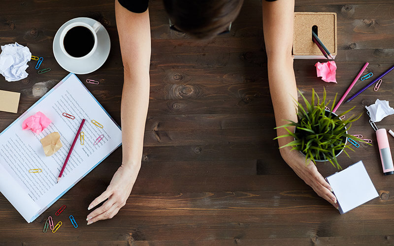 A woman attempts to clear her workspace by spreading her arms across her desk to part an assorted mess that includes paperclips, files, sticky notes, colored pencils, a cup of coffee and an office plant.