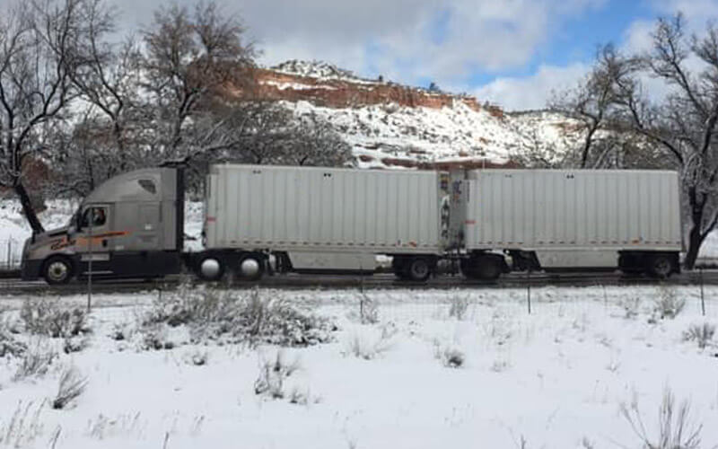 A grey Schneider semi-truck hauling two white trailers is parked at a rest area. There is snow on the ground a snow-covered mountain and trees in the background.