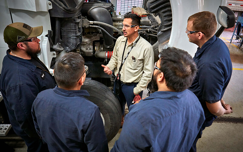 A group of four Schneider diesel technicians stand in a semi-circle around their training leader, who is talking, near a Schneider tractor.
