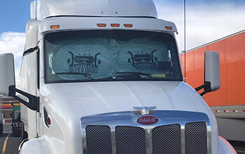 A white Peterbilt truck that is parked next to a Schneider trailer has a white sun shade with black lettering covering its front windshield.