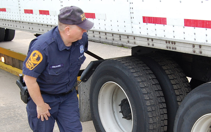 A uniformed officer inspecting the tires of a white tractor-trailer at a weigh station. The officer is wearing a dark blue uniform with a badge and patches.