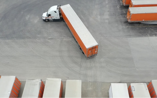 A white Schneider semi-truck hauling an orange trailer backs into a parking spot between other trailers.