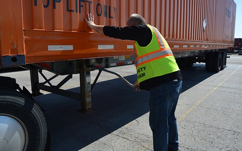 A driver wearing a black long sleeved shirt, safety vest and jeans cranks the landing gear of an Intermodal trailer.