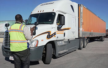 A training engineer wearing a tan shirt, neon vest and dark pants instructs a driving student as he backs a white semi-truck hauling an orange trailer in the truck yard.