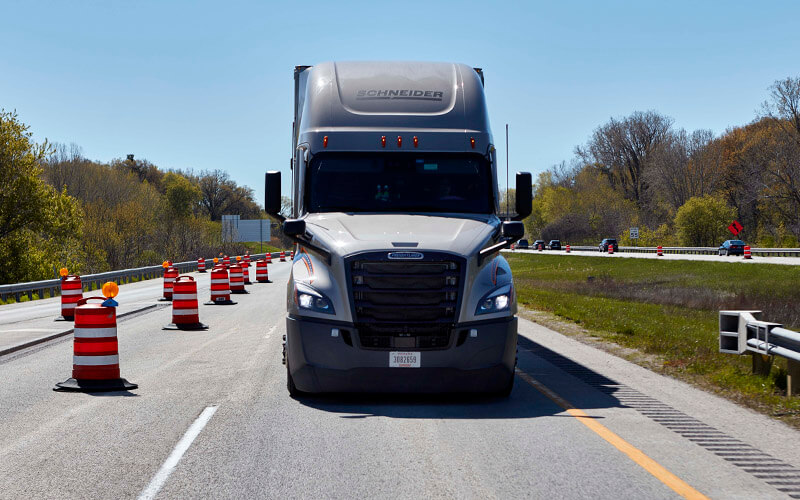 A grey Schneider semi-truck drives on a highway with road construction.