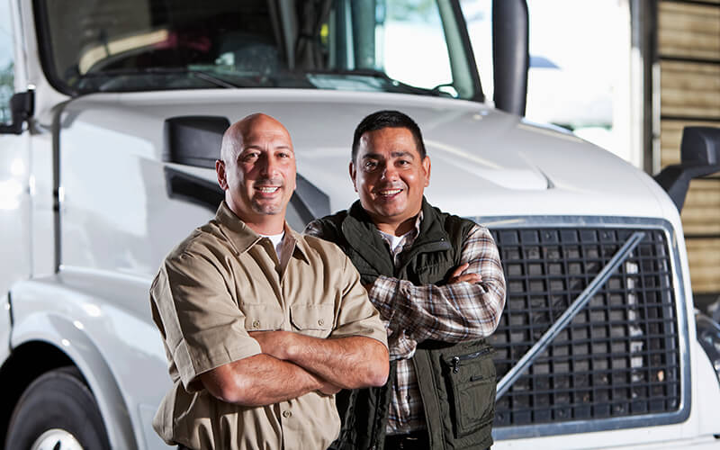 Two truck drivers pose with arms crossed in front of a truck.