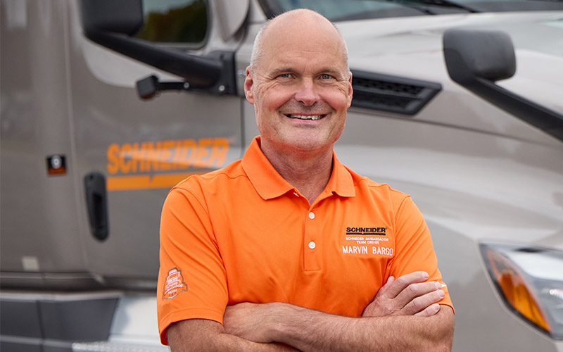 Marvin Bargo standing outside in front of a gray Schneider semi-truck with his arms crossed, smiling straight ahead.