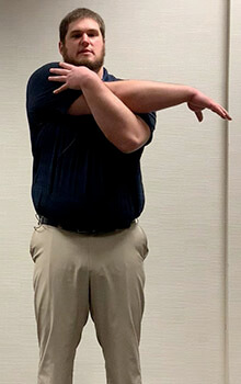 A physical therapist demonstrates stretching his shoulder by reaching is right arm across his body and grabbing his right shoulder with his left hand.