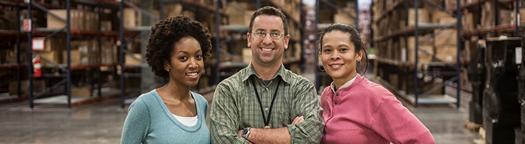 Three Schneider warehouse associates pose in front of several tall shelves of pallets and boxes