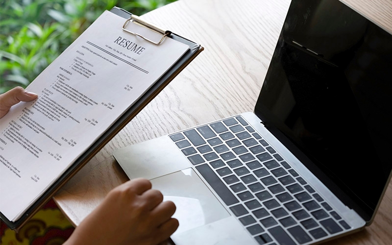 Someone holding a resume on a clipboard with a computer sitting on a table nearby.