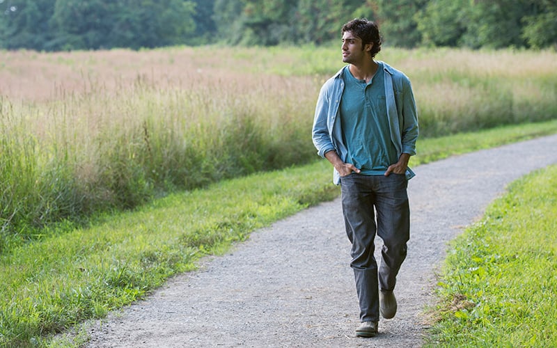 A truck driver walking along a gravel path through a grassy field bordered by trees.