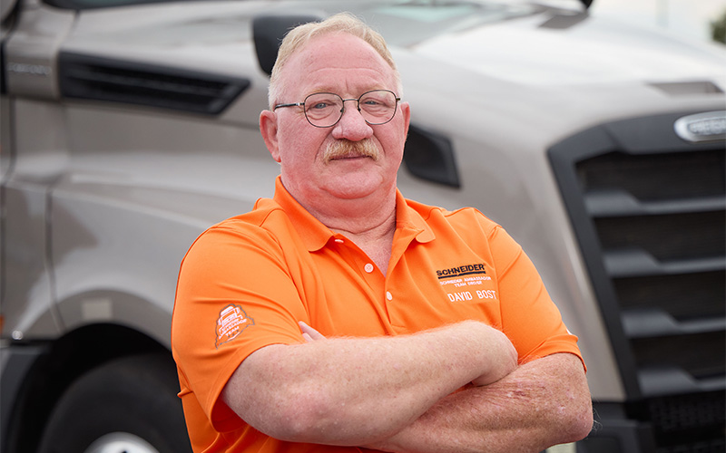 David Bost standing outside in front of a gray Schneider semi-truck with his arms crossed, looking straight ahead.