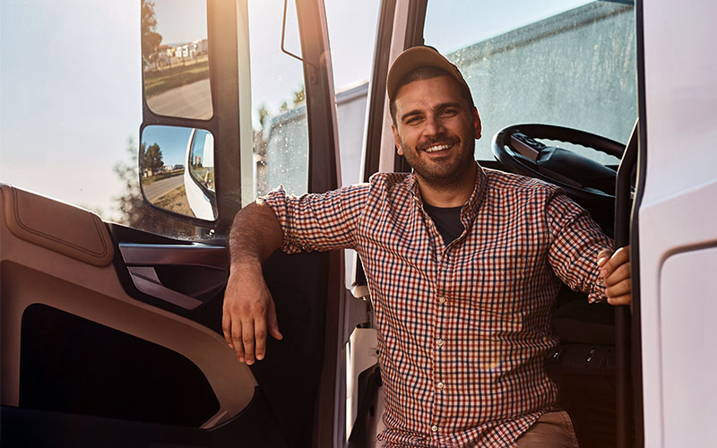 A truck driver stands between the open door of his truck and the cab.