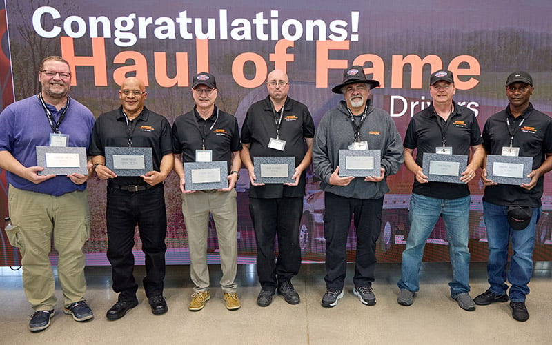 Seven drivers stand in front of a “Congratulations! Hall of Fame Drivers” display, holding recognition plaques during a driver appreciation event.