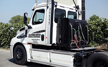 A white eCascadia truck is parked at an angle at a California facility. The tractor is not hooked up to a trailer, so the side batteries and plugs on the back of the truck are visible.