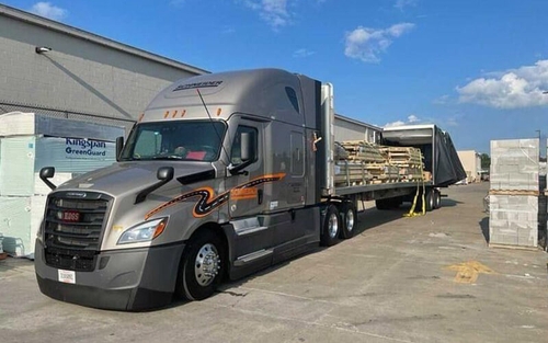 A grey Schneider semi-truck hauling a flatbed trailer is parked at a customer's loading area. The flatbed truck is loaded with wooden pallets that are strapped to the trailer.