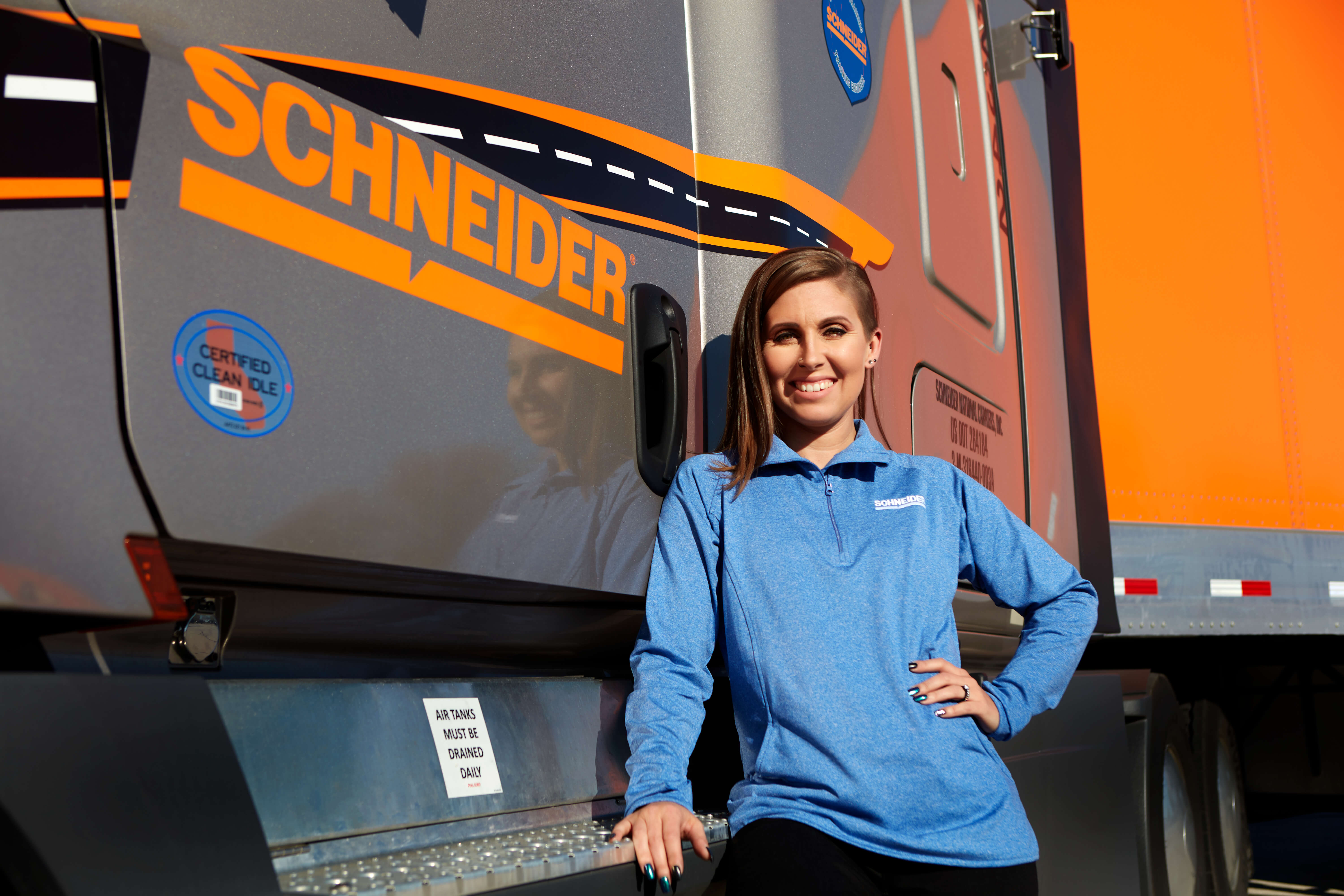 KayLeigh McCall poses in front of a champagne-colored Schneider tractor and an orange Schneider trailer.