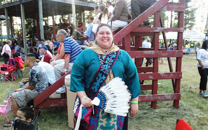 Kat wears her regalia at a Menominee gathering. The outfit includes a multi-colored patterned skirt, bandolier bag, long-sleeved top, scarf, bead necklaces and a feather fan.