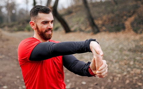 A truck driver wearing red and black performing a wrist stretch outside of their truck.