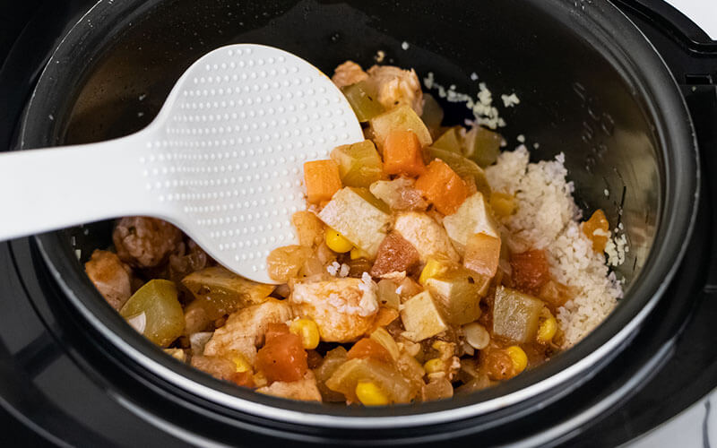 A white plastic spoon stirs a mixture of veggies, chicken and rice in a black crock pot.