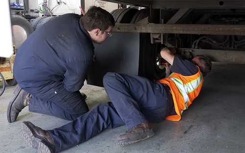 A Schneider diesel technician works on repairing the underside of a semi-trailer while his leader supervises.