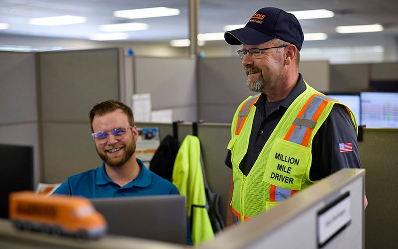 A truck driver wearing a high-visibility vest labeled “Million Mile Driver” stands beside a seated colleague at a cubicle workstation, with a laptop open and office partitions in the background.