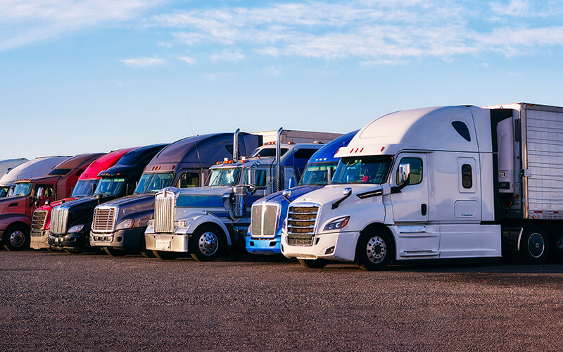 A variety of semi-trucks are parked side-by-side in a gravel parking lot.