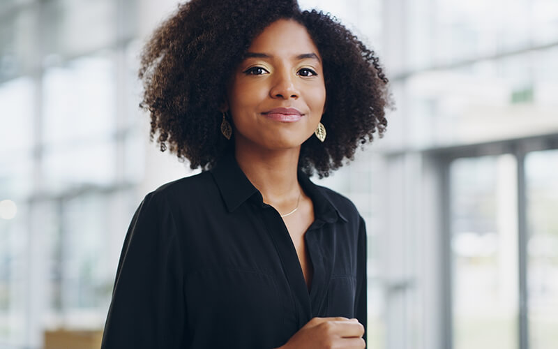 A young woman wearing a black collared shirt and a gold necklace and earrings, stands in front of a blurred out background.