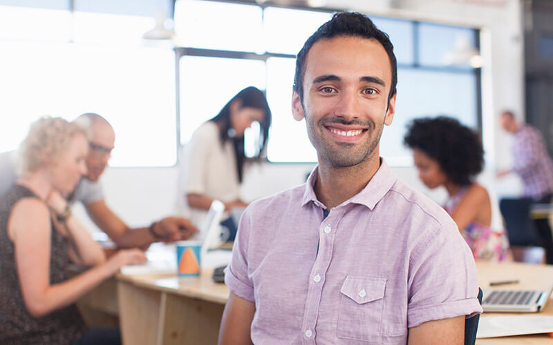 An office associate sits back in an office chair while colleagues collaborate at a conference table behind him.