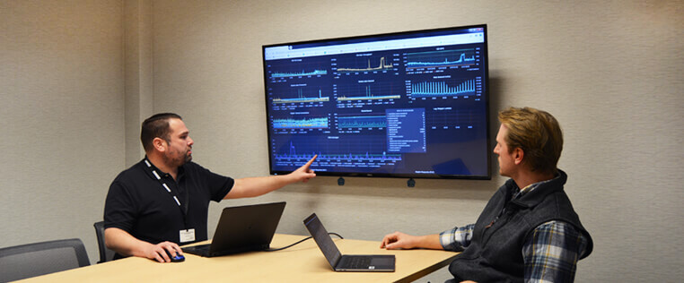 Two Schneider IT associates work at a table while one gestures at graphs on a large monitor nearby