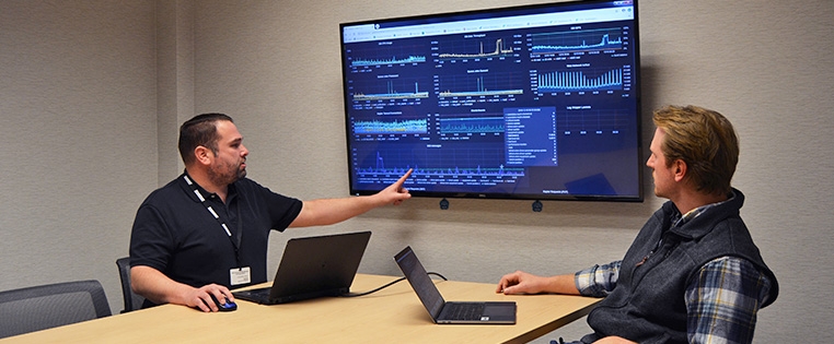 Two Schneider IT associates work at a table while one gestures at graphs on a large monitor nearby 