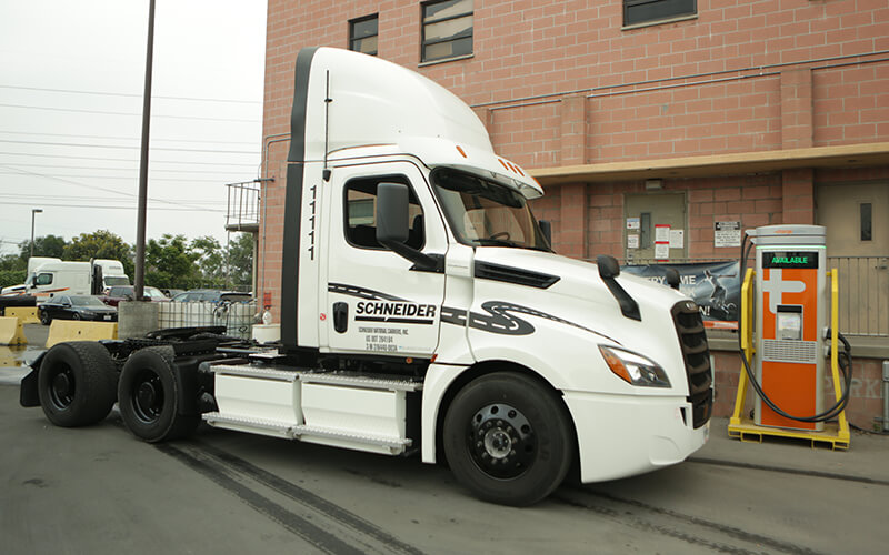 A white electric semi-tractor trailer is parked next to its charging station outside of one of Schneider’s California operating centers.