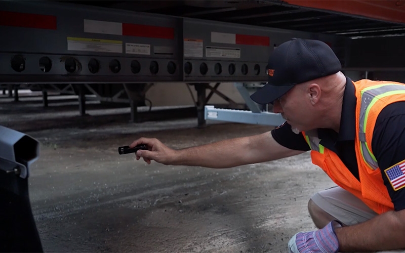 A truck driver wearing a black hat, shirt and neon orange safety vest uses a flashlight to inspect something under his semi-truck trailer.