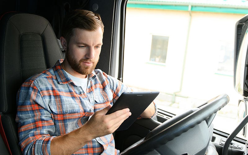 A person seated in the driver’s seat of a semi-truck cab holding and looking at a tablet device with both hands. 