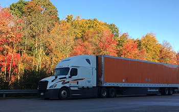 A Schneider tractor-trailer is parked alongside trees in the fall.