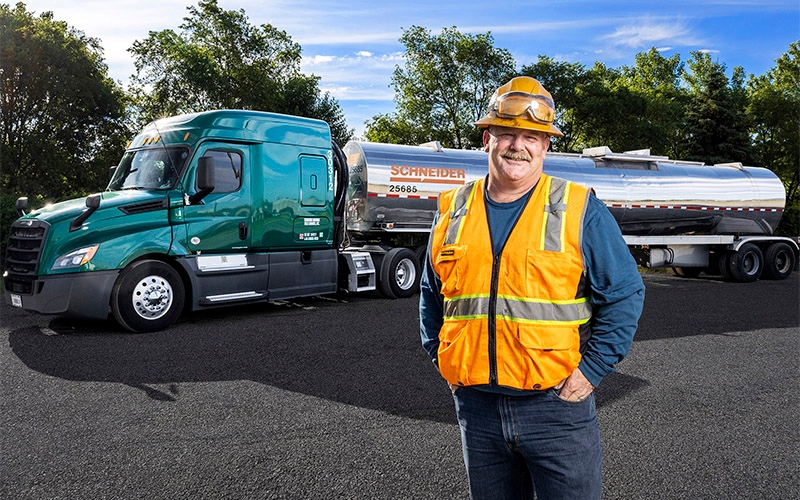 A Schneider tanker truck driver in orange PPE standing in front of a tanker trailer.