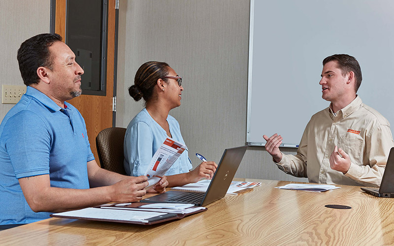 Three Schneider sales associates talk while seated around a table in a 
conference room with a whiteboard in the background