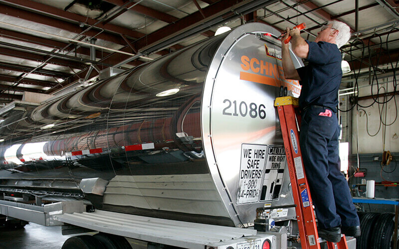 A Schneider diesel technician stands on a ladder to perform repair work on a company tanker trailer