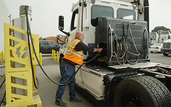 A bald truck driver wearing a safety vest, black shirt and jeans plugs the eCascadia semi-truck into the charging station.