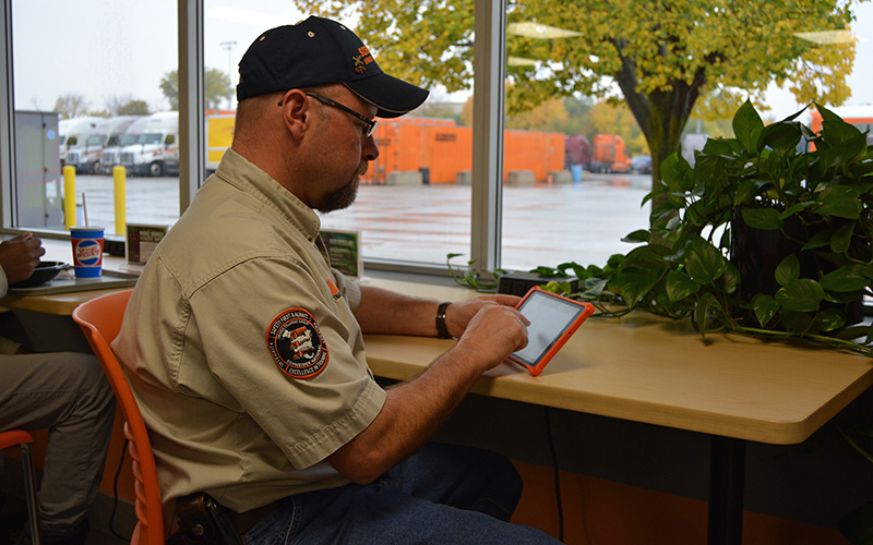 A Training Engineer wearing a tan shirt, black hat and glasses sits at an Operating Center cafeteria table and uses a driver tablet.