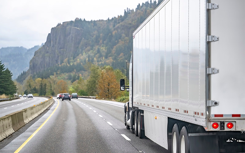 A white semi-truck traveling on the right lane of a highway. The trailer of the truck is visible, while the cab is not in view. The road is surrounded by lush greenery and trees, with a rocky hillside in the background.