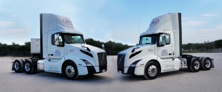 Two white Midwest Logistics Systems day cab semi trucks parked in a lot, facing nose to nose.