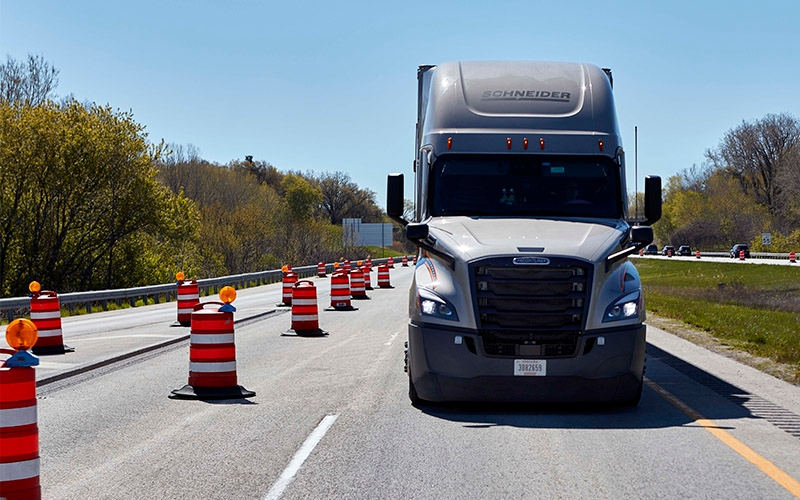 A grey Schneider semi-truck driving through a work zone lined with traffic barrels. 
