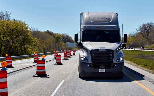 A grey Schneider semi-truck driving through a work zone lined with traffic barrels. 