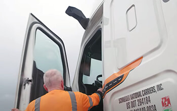 A driver wearing a reflective orange long sleeved shirt opens the door of a white Schneider semi-truck with MirrorEye cameras instead of standard mirrors.