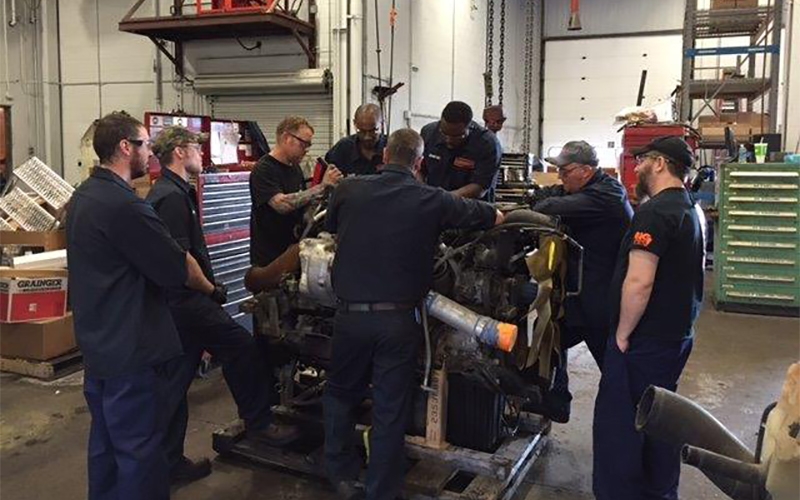 Schneider diesel technicians gather around a heap of truck components to watch an instructor perform a repair